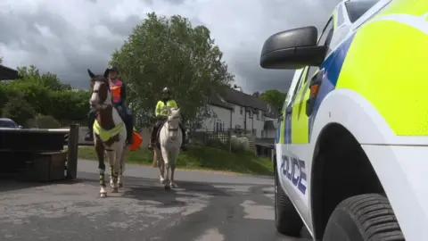 Two horses are seen approaching the camera with riders wearing high-viz jackets and a police car seen to the right. 