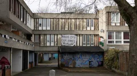 Facundo Arrizabalaga A banner reading “Hands Off Our Homes” hangs from a concrete housing block on the Lesnes Estate in Thamesmead. The ageing estate shows signs of wear, including peeling panels and graffiti, with a pedestrian walkway running beneath the building.