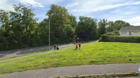 A general view of a green open space at the end of a road. An adult and child are walking across the grass. A person is on the pavement pushing a pushchair. Trees and a house are in the background.