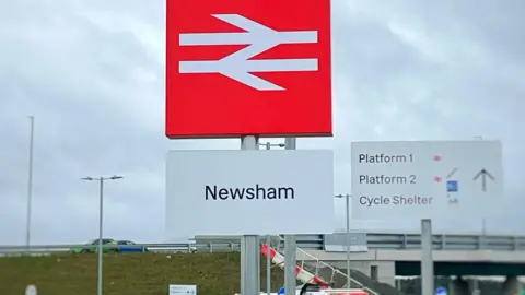 A sign saying Newsham is prominent with a red sign showing the British Rail logo in white above it. To one side is a sign saying Platform 1 and Platform 2. A bridge can be seen going across the line 