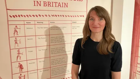 James Grant/BBC A woman in a black dress stands next to a wall chart showing different types of gladiators.