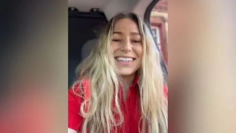 Sitting in a vehicle and wearing a red top, Katy Davies smiles at the camera. She has long blonde hair.