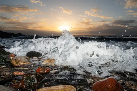Stephanie Virot Close-up of waves breaking over smooth shoreline stones at sunset, with water splashing into the air and the sun low on the horizon casting warm light through scattered clouds.