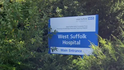 Google A Google Maps street view image of the entrance sign for West Suffolk Hospital. It is blue and white and set amongst some trees. It reads: West Suffolk Hospital Main Entrance
