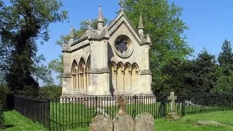 A stone-built mausoleum in a churchyard. It is in the shape of a small chapel and is surrounded by black railings. 