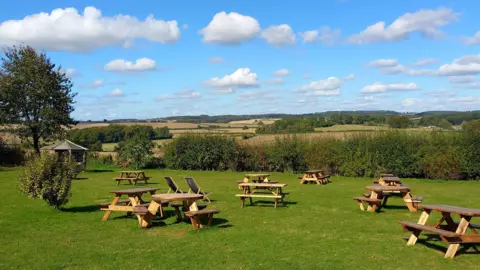 SteveC A lovely sunny day with picnic benches scattered on a grassy landscape looking out on to fields. The fields are lined with trees and hedges