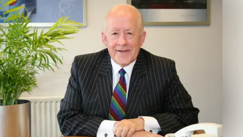 Alasdair MacConachie, who has a bald head and white hair at the sides and is wearing a black pin striped blazer, white shirt and colourful tie, sits with his hands crossed over one another at a desk. A landline phone is to his left and a plant to his right. 