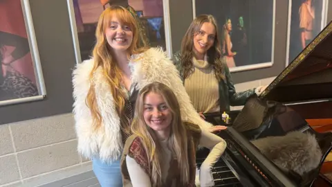 Lauren Byrne (left), Holly-Anne Hull (centre) and Charlotte Steele pose by a grand piano in a large room at a college. Holly-Anne is sitting on the piano stool and leaning on the piano while the other two stand behind her. The piano is black with an orange interior and several large photos of students are mounted on the wall behind them.