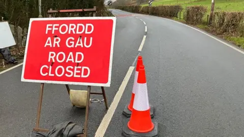 A red sign on BBC Road stating that it is closed. There are two cones on the right side of the sign. The road is in a rural area