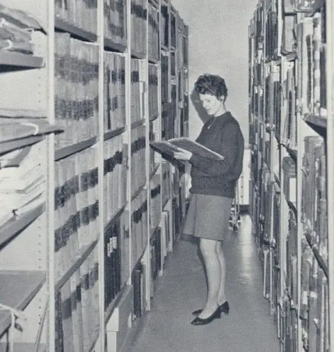 The Story/Durham County Council A woman stands between two large racks of folders. She wears 1960s-style clothing, with a short skirt and short hair. She is reading from a folder.