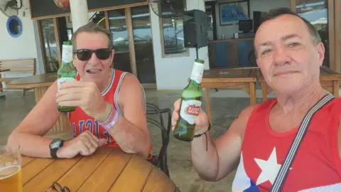 Mr Longworth, left, and Mr Alfonso, right, smile at the camera. They are both holding green bottles of Stella and wearing red vest tops with different writing on them. They are sitting at what appears to be an outdoors bar or restaurant. 