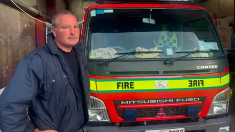 Siobhan Cox Finbar Gallagher is standing next to a red fire engine. The words fire is displayed across a green line on the vehicle. Gallagher has a navy jacket and jumper on. 