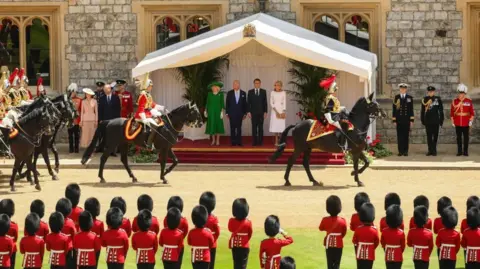 Reuters Queen Camilla, King Charles, French President Emmanuel Macron and his wife Brigitte stand in a white gazebo as they watch the Guard of Honour, made up of men dressed in red tunics and black bearskin caps, in the grounds of Windsor Castle. 