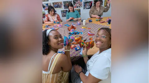 Think Human Five women sitting at a crafts table and making various colourful creations out of cloth and other materials. Two of them are turning back to the camera and smiling while holding their crafted items.