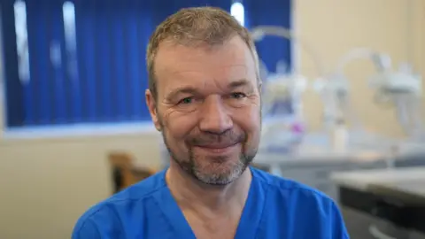 John Fairhall/ BBC Professor Nicholas Barker is wearing blue scrubs and looking at the lens of the camera, whilst smiling. The background which is of a dentistry training room is slightly blurred and machinery can be seen over his left shoulder.