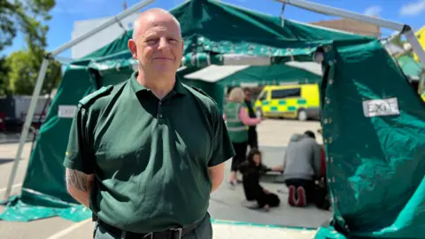 Jason smiles at the camera with his arms behind his back and in the background - a man is doing chest compressions on a dummy