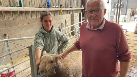 Luke Deal/BBC Amy Byford and Stephen Cobbald smile at the camera with a sheep stood between them. Mr Cobbald wears a red jumper with a shirt underneath and has glasses and grey hair. He rests one hand on the sheep's back.