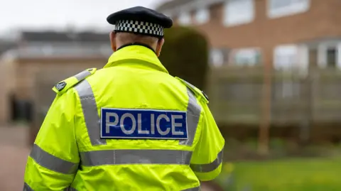 A stock image of a male police officer walking away from the camera towards brick houses. He is wearing a yellow hi-vis jacket which says POLICE across the back, and a black hat with a white chequered border. 