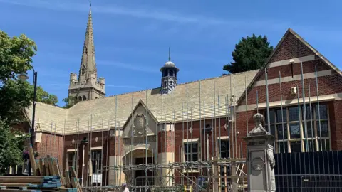 North Northamptonshire Council The outside of a large brick and stone building, with a church spire to the left, behind the building. There is scaffolding up outside the building. There is also a large blue sky. Trees are to the left and behind the building to the right. 