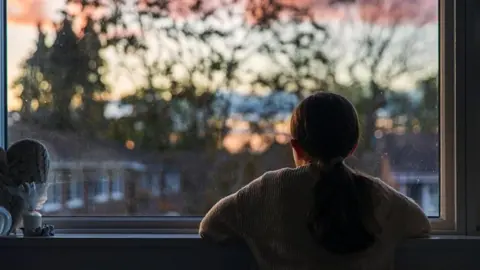 Getty Images A young girl stares out of a window at dusk, with her elbows on the window ledge. There are properties in front with a backdrop of tall trees.