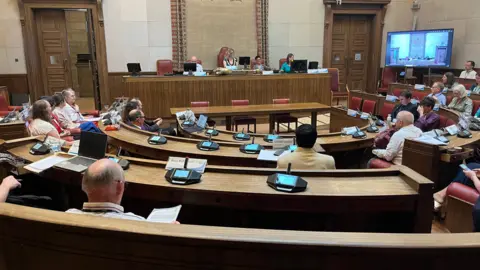 Phil Shepka/BBC A view of a council meeting. Councillors sit in semi-circular rows in a large chamber. 