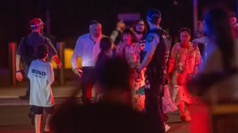 EPA A group of people walk towards a police officer. It is dark, and the people are lit by red light. The police officer has his back to the camera, two women are wearing summer dresses, while a man is dressed in a shirt and trousers, and wearing a kippah. Another boy walks away, wearing a white t-shirt with Bondi written on it