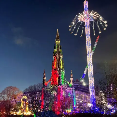 Duncan MacPherson The Scott Monument in Edinburgh is in the middle of the image. It is dark with green and red lights on either side. A large fairground ride is to the right and is lit up in white and blue. A smaller ride is to its left and is lit up in yellow.