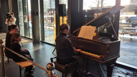 A young girl plays a piano. Two other girls are sat behind her, watching. A TV camera is placed on the side, with a person operating it. The brown wooden piano is placed in front of a glass-fronted wall.