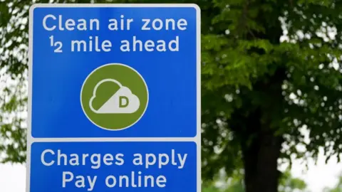 A bright blue sign with a green circular logo containing a white cloud with a green letter D inside it. Above the logo is white text over two lines reading: "Clean air zone 1/2 mile ahead". Below the logo is a separate blue box with white text over two lines reading: "Charges apply Pay online". A tree is in the blurred background
