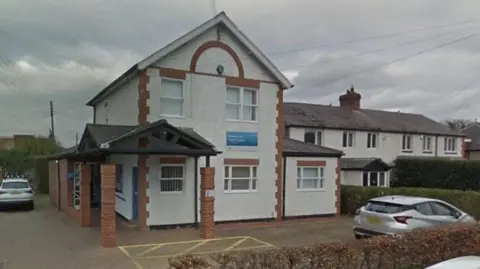 Rowlands Gill Medical Centre. The building is a white converted house with some red bricks on its corners. Two cars are parked in a small car park outside.