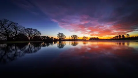 There is a large expanse of water with trees lining its distant shore. The trees are silhouetted against the sky, which is a dark blue colour but also bright orange on the horizon and peppered with pink clouds.