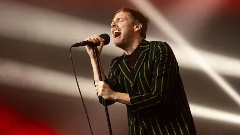 Ricky Wilson of Kaiser Chiefs singing into a microphone on a stand. He is wearing a black suit with bright green pinstripes.