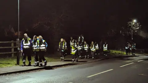 A group of young people wearing reflective gear walk on a pavement on the side of a road at night.