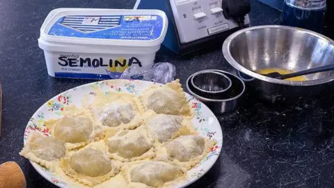 John Reed A plate is full of hand made ravioli made by one of the 2024 contestants. There is a box marked semolina and a mixing bowl with an egg mixture and spatula in it, beside some cutting shapes.