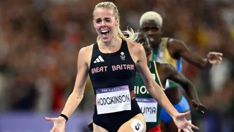 Keely Hodgkinson runs over the finish line at the 2024 Paris Olympics. She has her hands outstretched and has a look of utter joy on her face. 