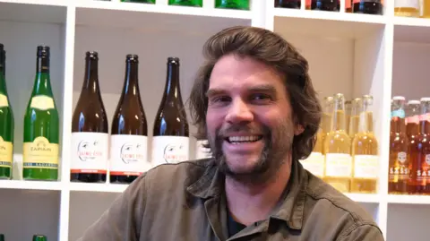 A man sits in front of a white shelf that is lined with bottles of cider. He has flowing dark hair with a large handle bar moustache.