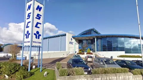 Google The National Sports Centre is a blue and white building with a wall of glass panes on the right and a sign tower to the front left reading: NSC. There are cars parked in front on a sunny day.