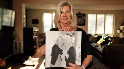 Christine, holding a picture of her with her mother, Dorothy Readhead. Christine has straight, shoulder-length blonde hair and is wearing a black long-sleeved top. She is pictured in a living room with windows and sofas behind her. In the picture she is holding, she has her arms round her mother, and they are laughing and cuddling each other.