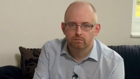 Chris Whitaker, with short fair hair and a close cropped beard, wearing rectangular steel-rimmed glasses, and a light blue shirt, sitting on a dark sofa looking serious and straight at the camera