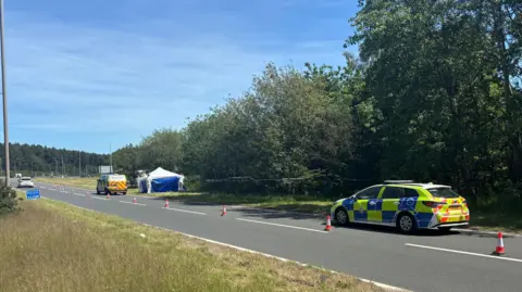 BBC A blue and white forensics tent next to two police cars and cones blocking off a lane of a highway