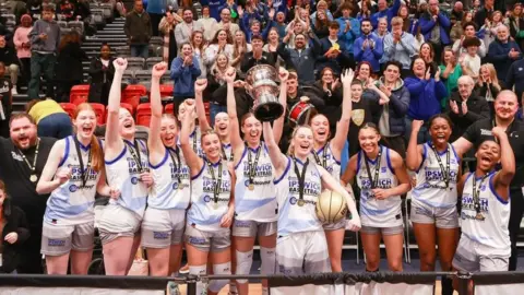 Twelve female Ipswich basketball players dressed in a white kit with a blue trim lift a silver trophy in front of applauding fans.