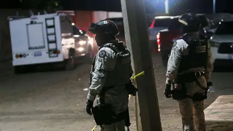 Members of the National Guard stand at the crime scene where at least 11 people were killed and 12 more wounded during an armed attack at a football field in Salamanca, Guanajuato state, Mexico, on January 25, 2026. The two men are wearing helmets and are in uniform. Cars and vans can be seen in the background as well as yellow crime scene tape. 
