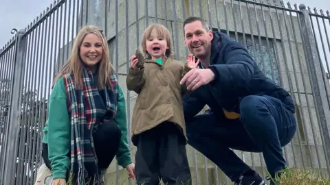 The Harte family with their young daughter Grainne at the base of the St Patrick's monument on Slieve Patrick.    Her mother Maeve has shoulder-length straight blonde hair and is wearing a green sweatshirt and a read, white and blue tartan scarf.  Grainne has short blonde hair, a beige overcoat and is holding a pine cone. Her father has short, greying hair and is wearing a navy coat and dark blue jeans. 