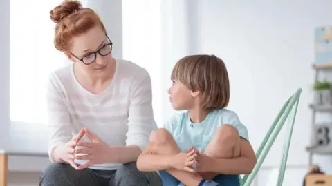Getty Images An adult wearing glasses and a white top sits facing a child seated on a chair in a bright indoor room. The child is sitting with knees drawn up and hands clasped around the legs, while both look towards each other. The background is softly blurred, with shelves and household items visible.