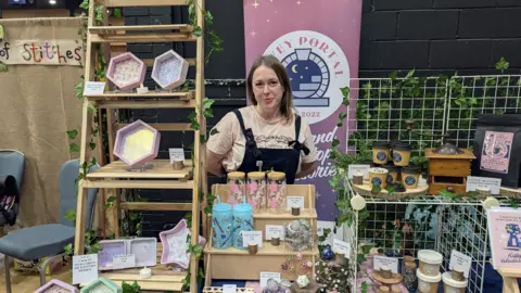 Angie Taylor, a blonde woman in a T-shirt and dungarees, stands behind her stall. It includes jewellery, coffee glasses and dice trays. 