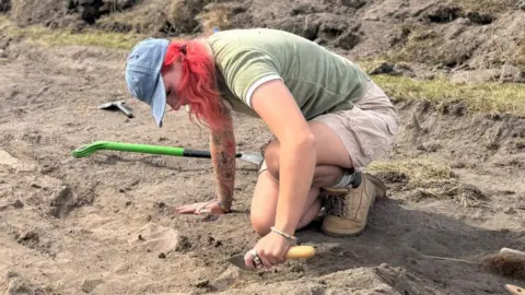 Supplied Meg Russell wearing a blue cap, green shirt, beige shorts, and work boots kneels in an archaeological excavation trench, carefully using a hand trowel to uncover features in sandy soil. Digging tools and spoil heaps surround the excavation area.