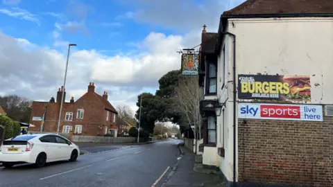 A white car drives down a road infront of a derelict pub