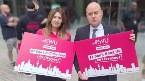 Carl Webb CWU A woman with long dark blonde hair and a bald man stand in front of an office building looking serious. She is wearing a pink shirt, he is wearing a white shirt with dark tie. They are holding bright pink placards which say "BT Don't Pull the Plug on Liverpool". 