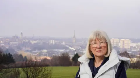 LDRS Wakefield councillor Maureen Cummings, a woman with white shoulder length hair. She is posing for a photograph on a hill, with the Wakefield skyline behind her.