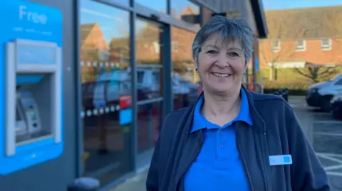 Simon Spark/BBC Co-op worker Abi Hill stands outside the glass store entrance. She has short dark hair and is smiling. She wears a Co-op blue polo shirt and fleece with a name badge.